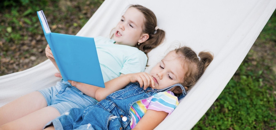 Two children lying in a hammock, one reading a book outdoors