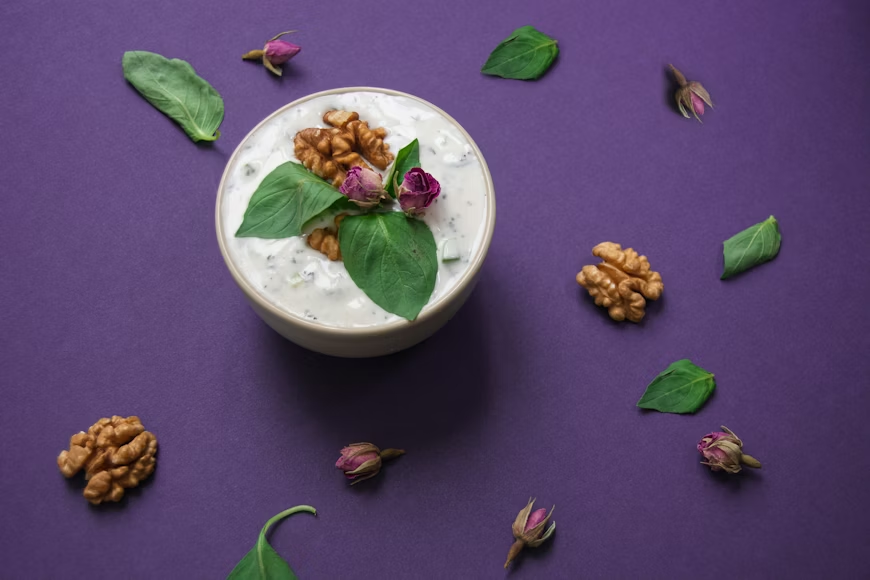 Bowl of yogurt topped with walnuts, green leaves, and pink rosebuds on a purple background, surrounded by more walnuts, leaves, and rosebuds.
