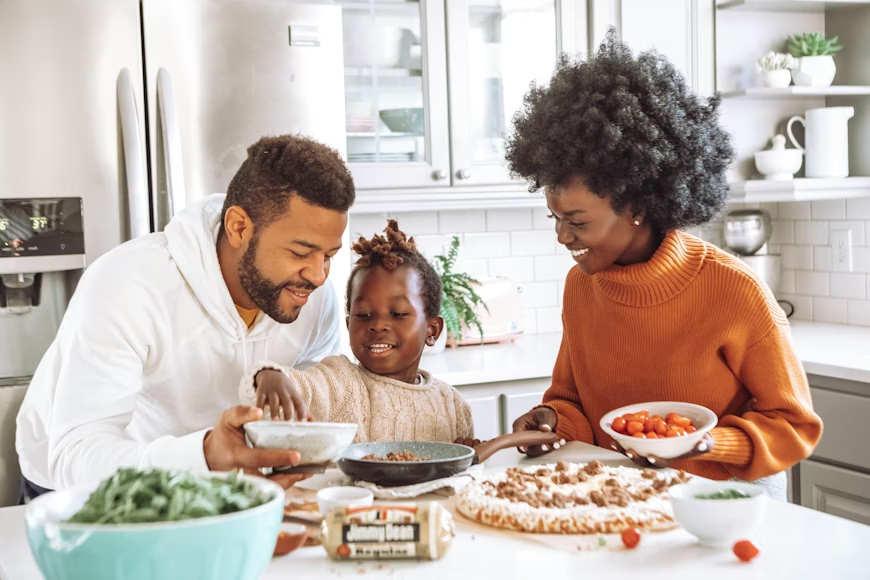 A joyful family cooks together in a bright kitchen while adding toppings to a pizza.