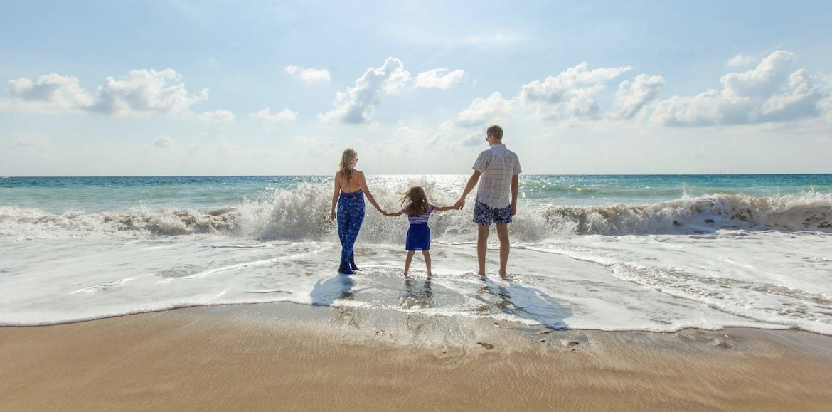 A family of three—a man, a woman, and a young child—standing hand-in-hand on a sandy beach, facing the turquoise ocean waves under a bright, sunny sky.