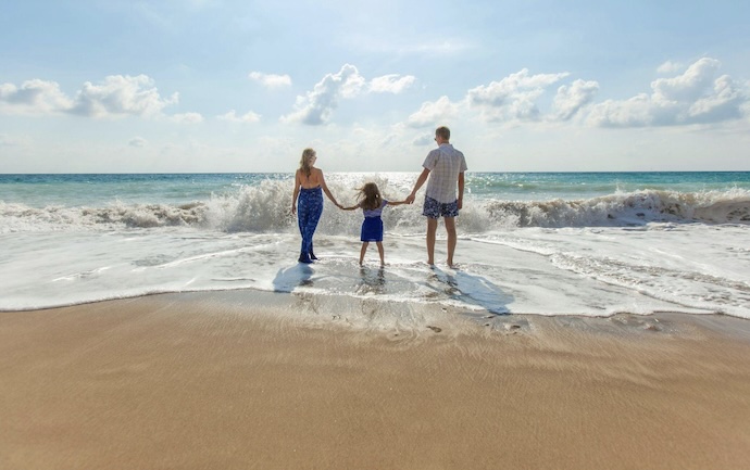 A family of three—a man, a woman, and a young child—standing hand-in-hand on a sandy beach, facing the turquoise ocean waves under a bright, sunny sky.