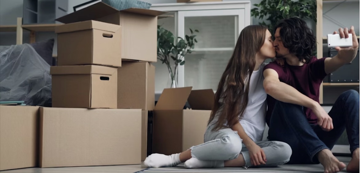 A couple kissing and clicking a selfie with their phone, with packed boxes placed beside them. 