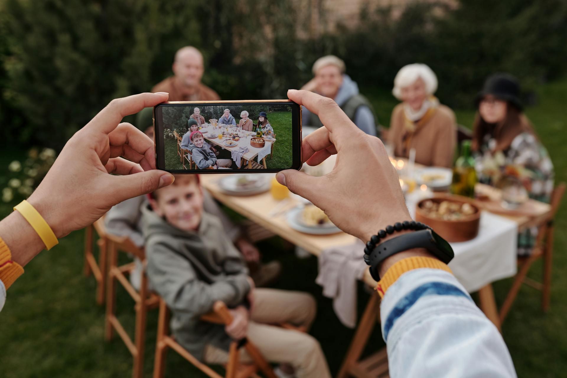 Hands holding a smartphone to take a photo of a multi-generational family enjoying a meal together at an outdoor table
