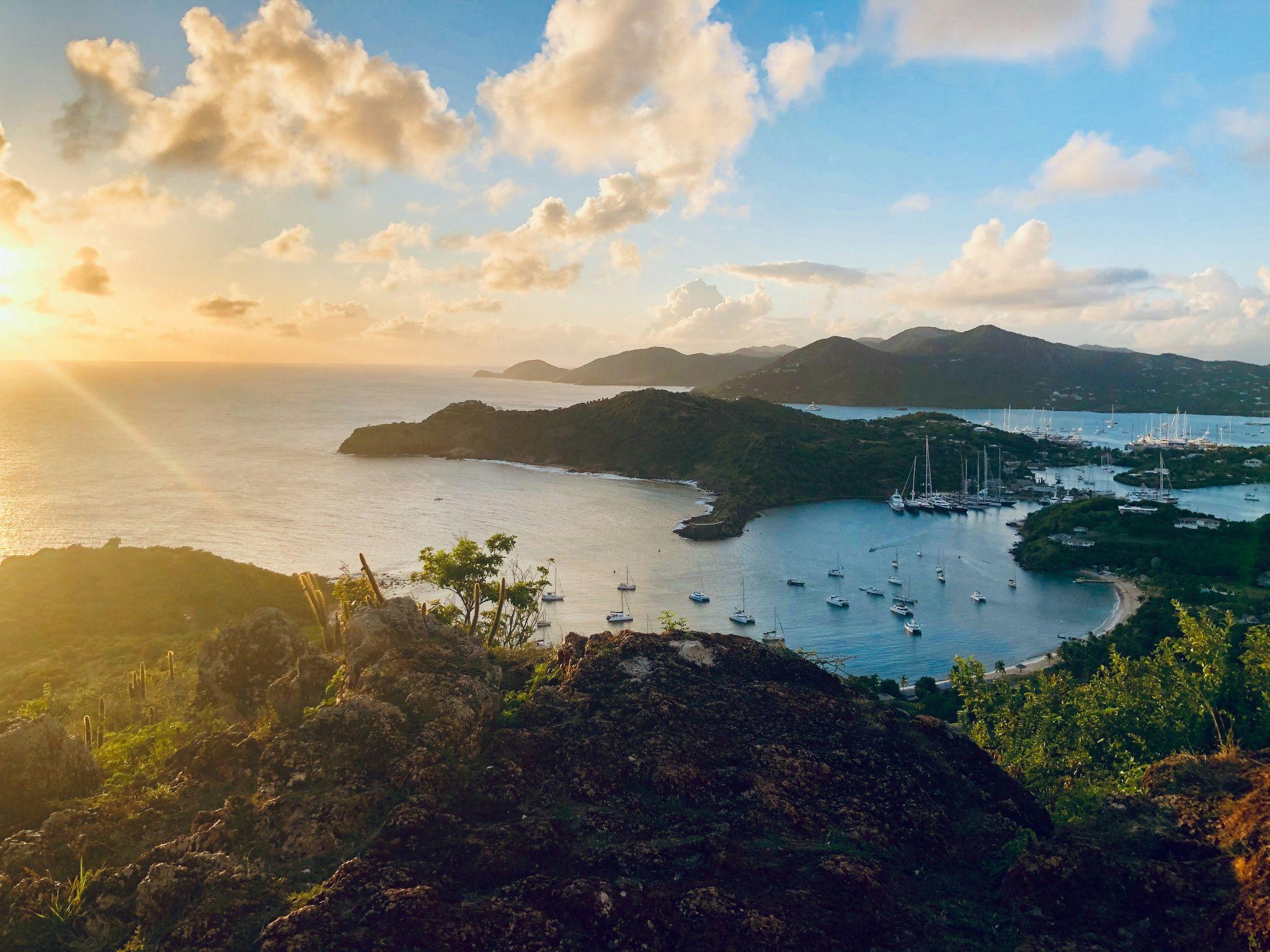 A scenic golden hour view of a coastal bay filled with sailboats, seen from a rocky hilltop overlooking lush islands and the glowing ocean at sunset.