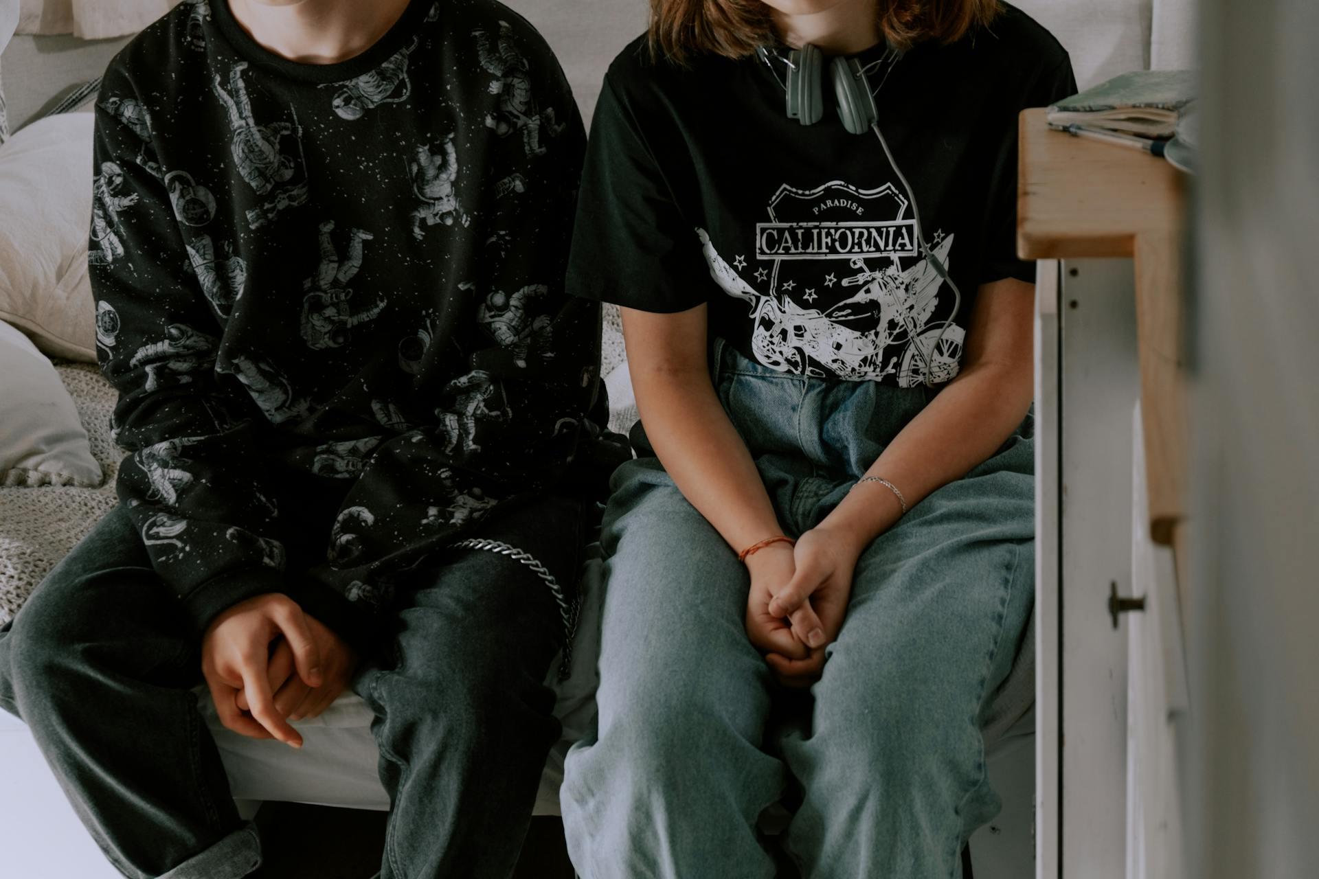 Two teenagers sitting closely together on a bed wearing casual dark clothing.