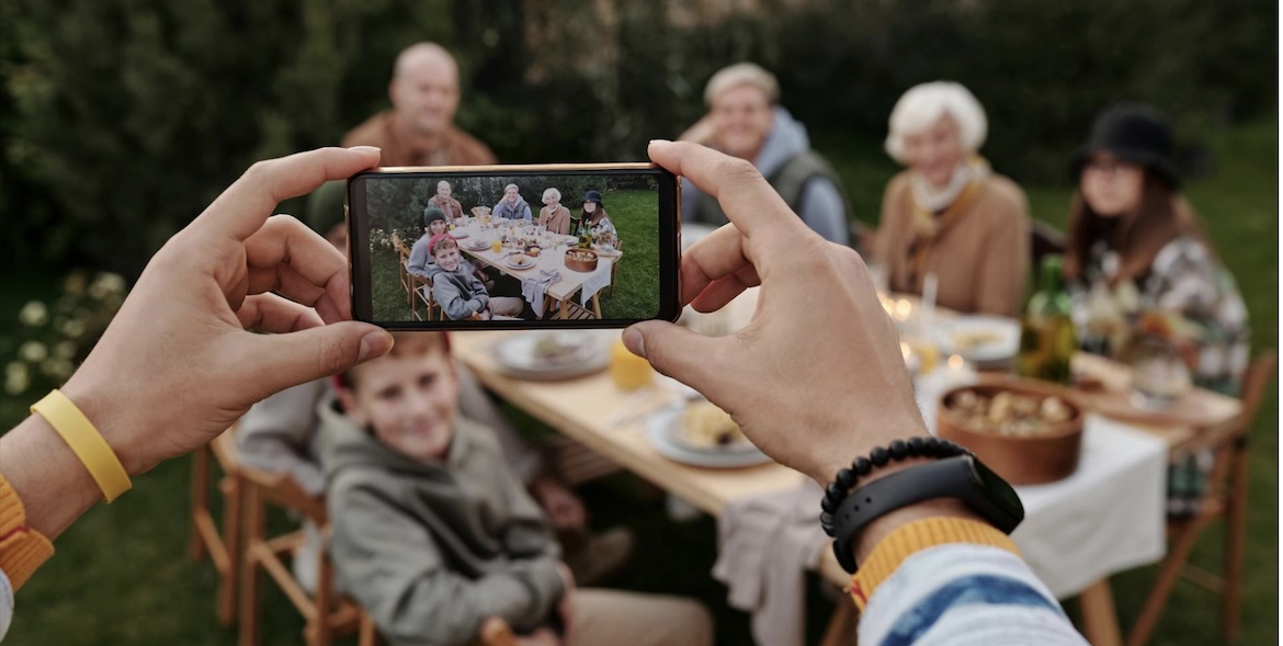 Hands holding a smartphone to take a photo of a multi-generational family enjoying a meal together at an outdoor table