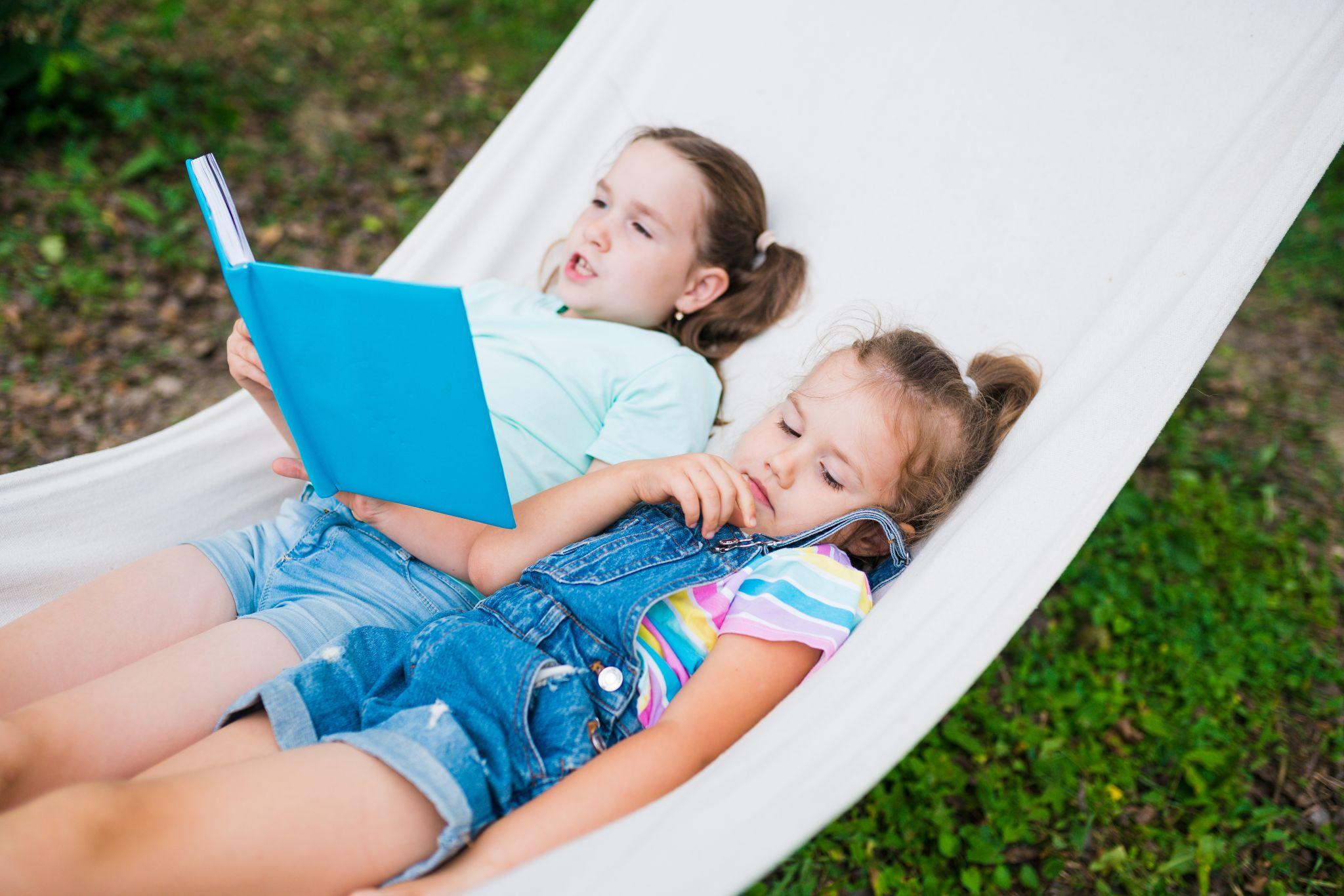 Two children lying in a hammock, one reading a book outdoors