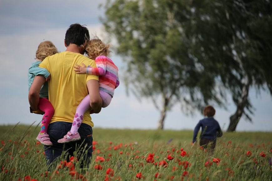 A man carries two children through a field of red flowers, with another child running ahead.