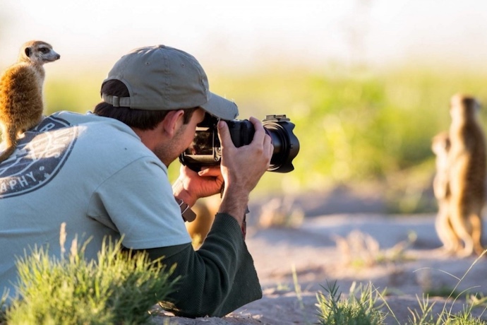 a man takes a photo of a meerkat whilst a meerkat sits on his back