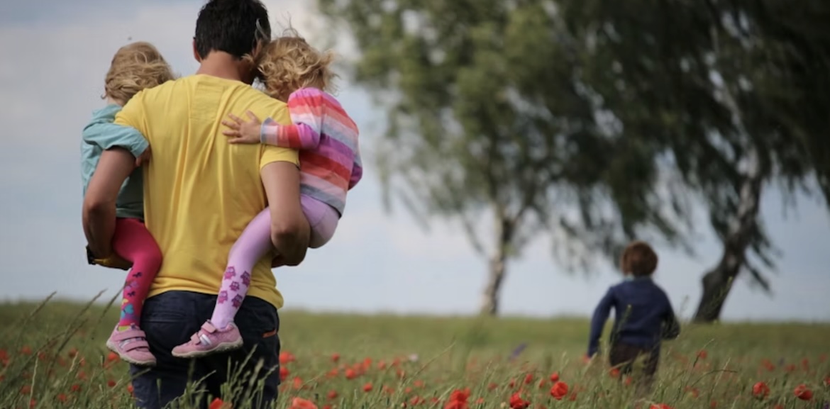  A man carries two children through a field of red flowers, with another child running ahead.