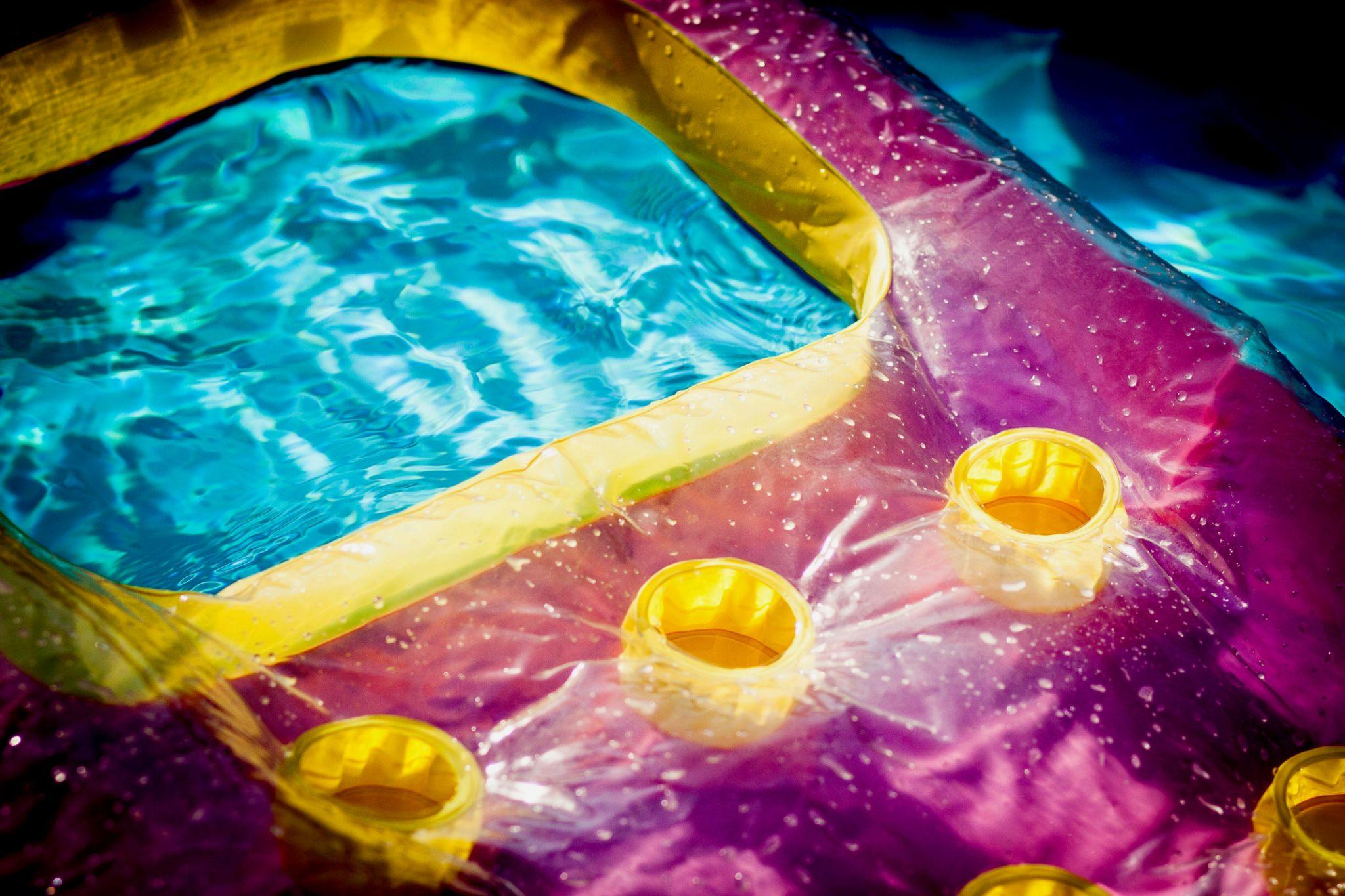 A close-up, high-angle view of a vibrant purple and yellow inflatable pool float drifting on clear blue water, with sunlight creating bright reflections on the rippled surface.