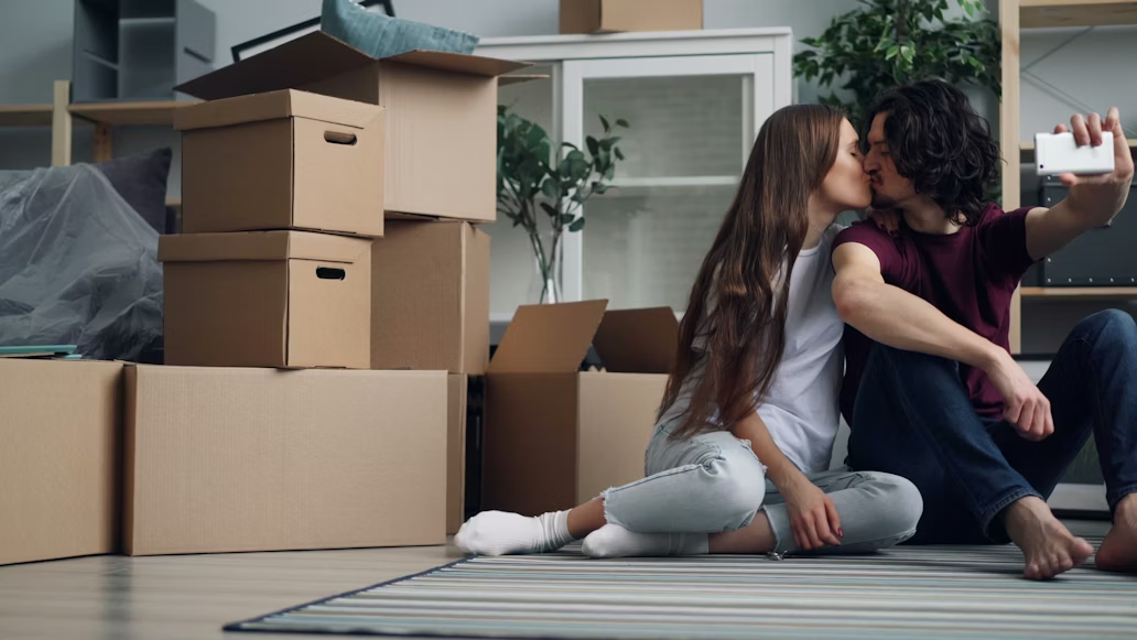 A couple kissing and clicking a selfie with their phone, with packed boxes placed beside them.