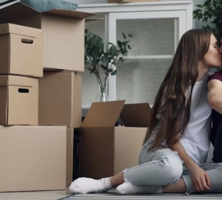A couple kissing and clicking a selfie with their phone, with packed boxes placed beside them. 