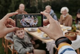 Hands holding a smartphone to take a photo of a multi-generational family enjoying a meal together at an outdoor table