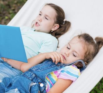 Two children lying in a hammock, one reading a book outdoors