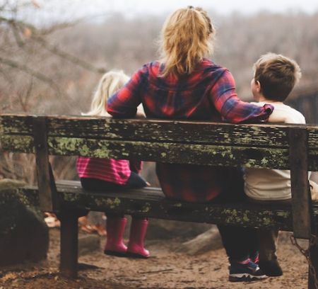 a mum and two kids sit on a bench in nature