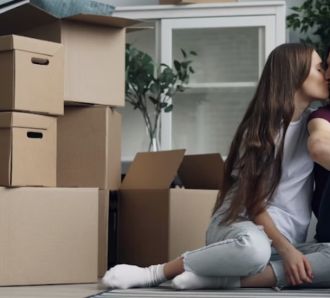 A couple kissing and clicking a selfie with their phone, with packed boxes placed beside them. 
