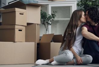 A couple kissing and clicking a selfie with their phone, with packed boxes placed beside them. 
