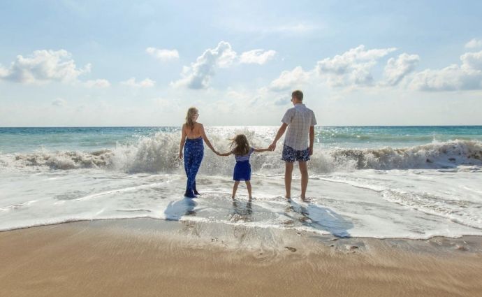 A family of three—a man, a woman, and a young child—standing hand-in-hand on a sandy beach, facing the turquoise ocean waves under a bright, sunny sky.