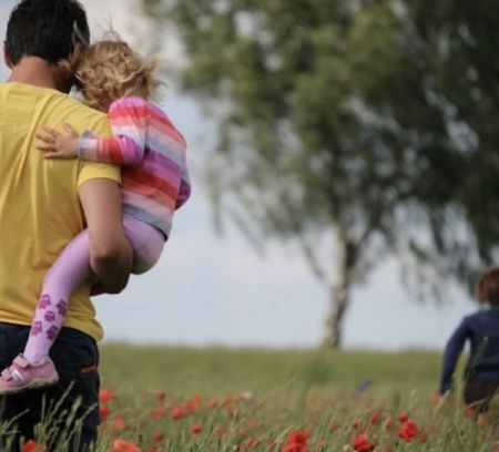  A man carries two children through a field of red flowers, with another child running ahead.