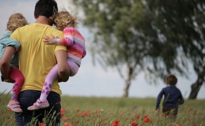  A man carries two children through a field of red flowers, with another child running ahead.