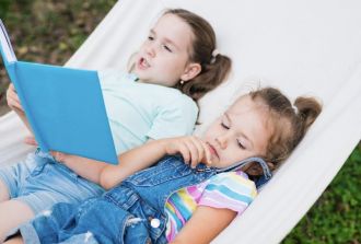 Two children lying in a hammock, one reading a book outdoors